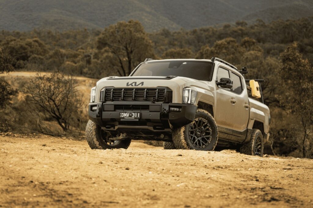 A beige Kia Tasman ute driving on a dry, dusty dirt road in a scrubby Australian environment